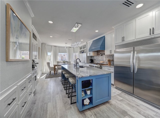 kitchen featuring premium range hood, white cabinetry, an island with sink, ornamental molding, and stainless steel built in fridge