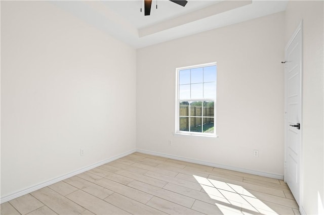 empty room with a tray ceiling, light wood-type flooring, a ceiling fan, and baseboards