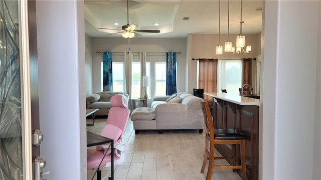 living room featuring light wood-type flooring, a tray ceiling, sink, and ceiling fan with notable chandelier