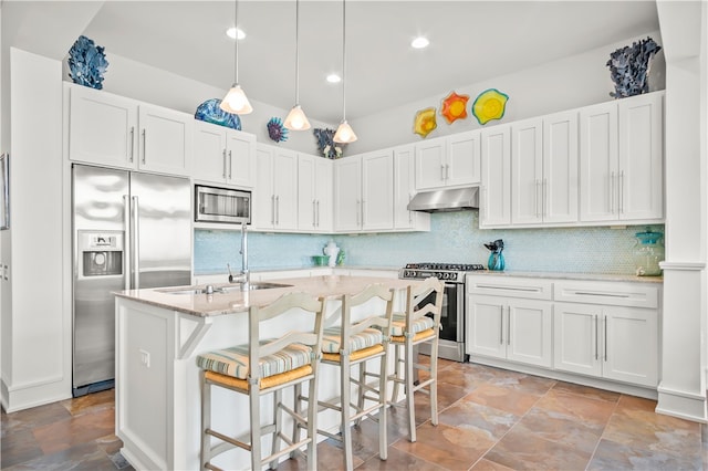 kitchen featuring built in appliances, a breakfast bar area, decorative light fixtures, an island with sink, and white cabinets