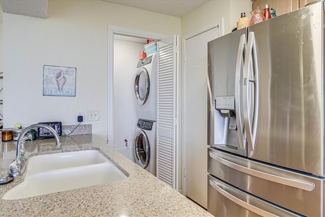 kitchen featuring a textured ceiling, sink, stainless steel fridge with ice dispenser, stacked washer / dryer, and light stone countertops