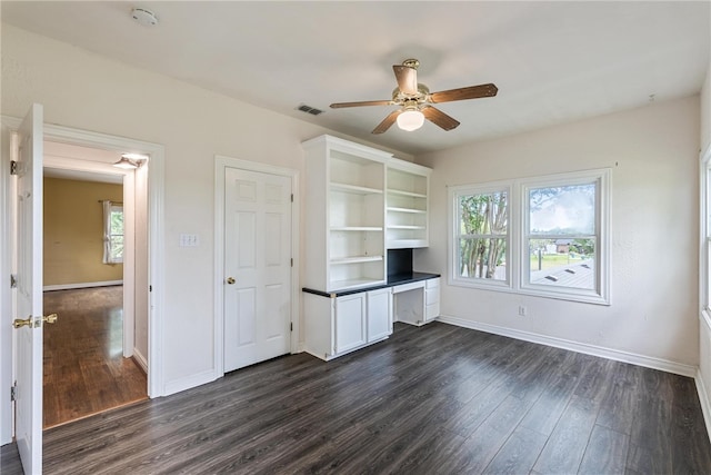 unfurnished office featuring a healthy amount of sunlight, dark hardwood / wood-style flooring, built in desk, and ceiling fan