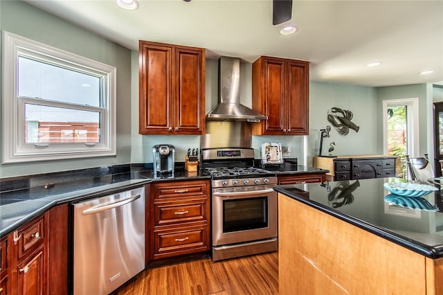 kitchen featuring dark stone counters, light hardwood / wood-style floors, wall chimney exhaust hood, and appliances with stainless steel finishes