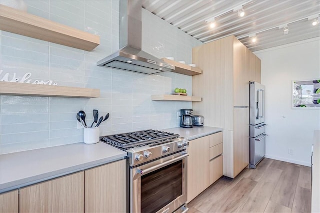 kitchen featuring light brown cabinetry, backsplash, stainless steel appliances, wall chimney range hood, and light wood-type flooring