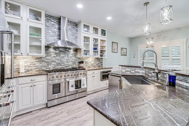 kitchen featuring appliances with stainless steel finishes, dark stone counters, wall chimney range hood, decorative light fixtures, and white cabinetry