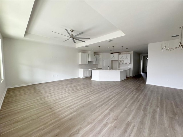 unfurnished living room featuring light hardwood / wood-style flooring, sink, a tray ceiling, and ceiling fan with notable chandelier