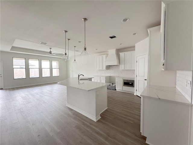 kitchen with custom exhaust hood, white cabinetry, light stone counters, a center island with sink, and pendant lighting