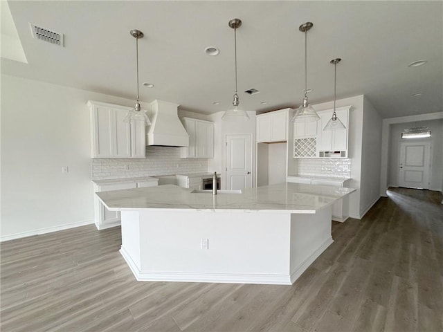 kitchen featuring custom exhaust hood, wood-type flooring, hanging light fixtures, a large island with sink, and white cabinets