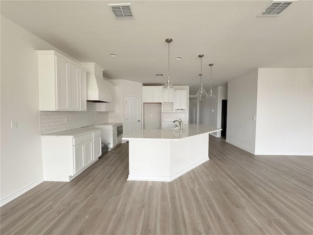 kitchen with custom exhaust hood, light wood-type flooring, a center island with sink, and white cabinets