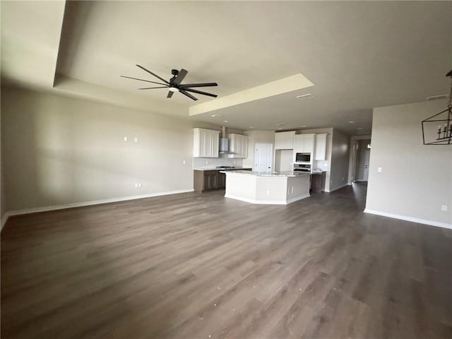 unfurnished living room featuring a ceiling fan, a tray ceiling, baseboards, and dark wood-style floors