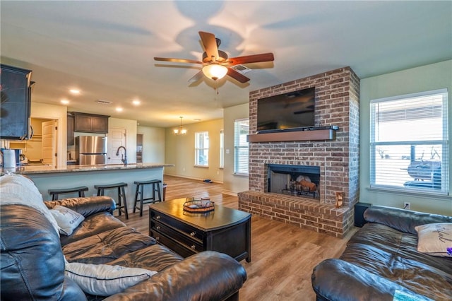 living room with ceiling fan with notable chandelier, a fireplace, sink, and light wood-type flooring