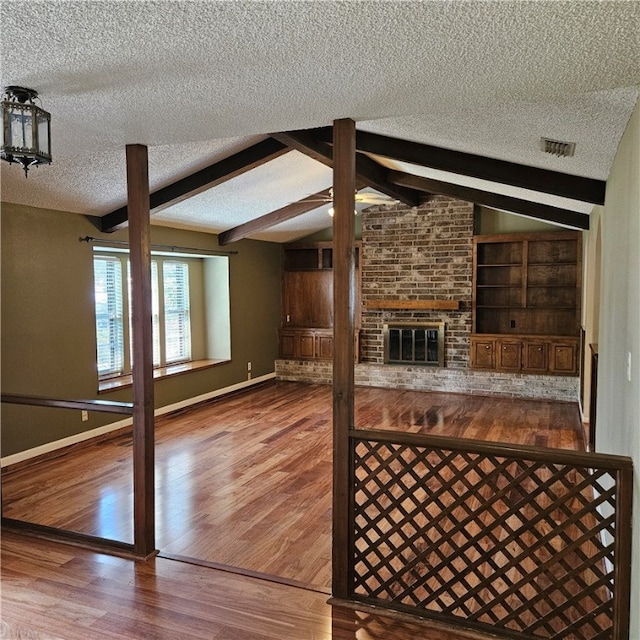 unfurnished living room with built in shelves, a textured ceiling, hardwood / wood-style floors, vaulted ceiling with beams, and a brick fireplace