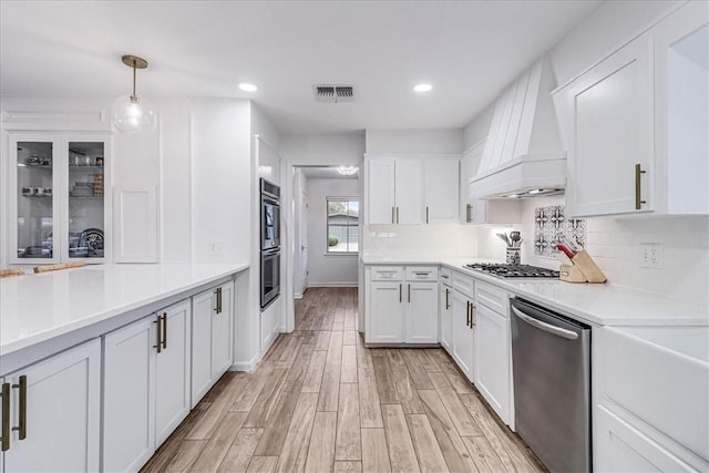 kitchen featuring custom exhaust hood, white cabinetry, hanging light fixtures, light wood-type flooring, and appliances with stainless steel finishes