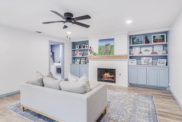 living room featuring ceiling fan, a brick fireplace, washer / clothes dryer, light wood-type flooring, and built in shelves