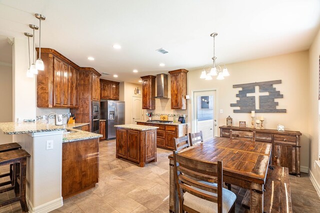 kitchen featuring stainless steel appliances, kitchen peninsula, light stone countertops, decorative light fixtures, and wall chimney range hood