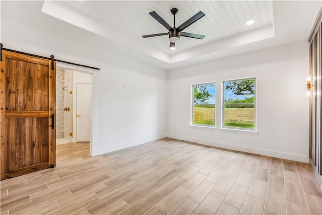 unfurnished bedroom featuring ceiling fan, a barn door, a raised ceiling, baseboards, and light wood finished floors