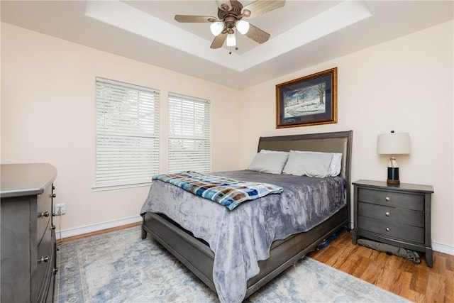 bedroom featuring ceiling fan, a raised ceiling, light wood-style flooring, and baseboards