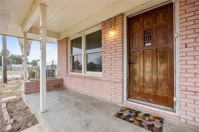 doorway to property featuring covered porch, brick siding, and fence