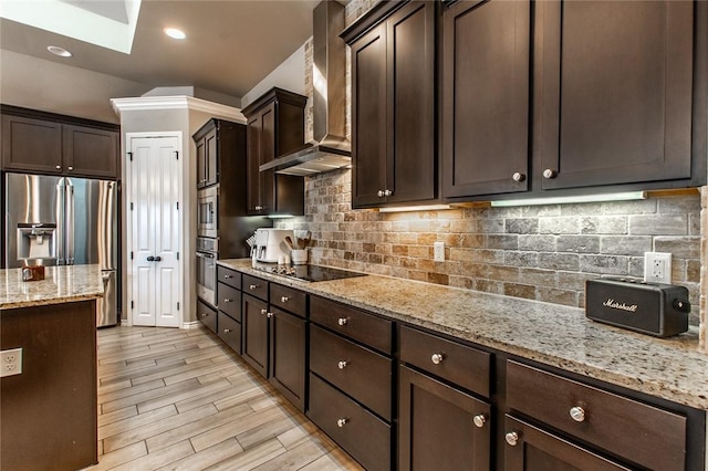 kitchen featuring wall chimney range hood, appliances with stainless steel finishes, dark brown cabinets, light stone counters, and light hardwood / wood-style floors