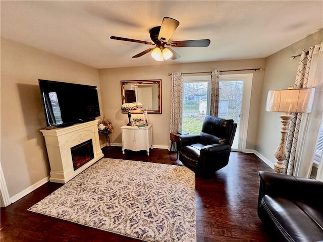 living area with a ceiling fan, dark wood-style flooring, a warm lit fireplace, and baseboards