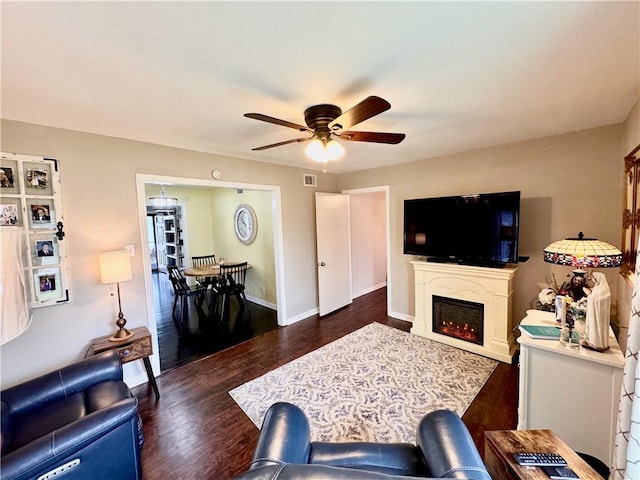 living area with dark wood-style floors, visible vents, ceiling fan, a warm lit fireplace, and baseboards