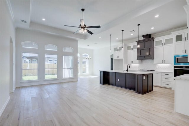 kitchen featuring stainless steel microwave, an island with sink, black oven, decorative backsplash, and ceiling fan with notable chandelier