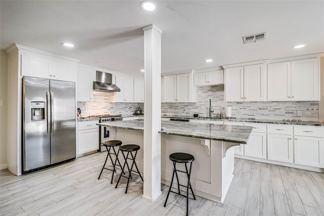 kitchen with stainless steel appliances, a breakfast bar area, wall chimney exhaust hood, and light stone counters