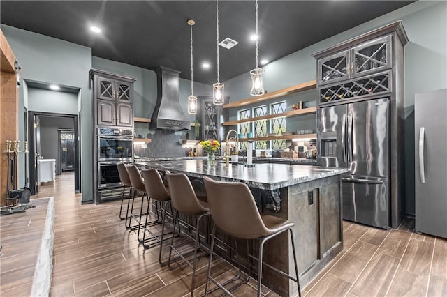 kitchen featuring wall chimney range hood, dark hardwood / wood-style floors, pendant lighting, a center island with sink, and appliances with stainless steel finishes