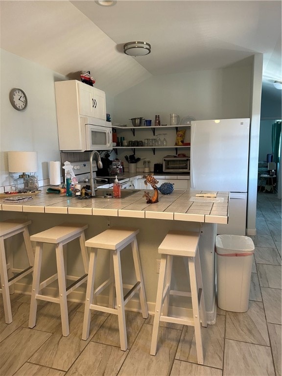 kitchen with tile counters, kitchen peninsula, a breakfast bar area, white cabinetry, and white appliances