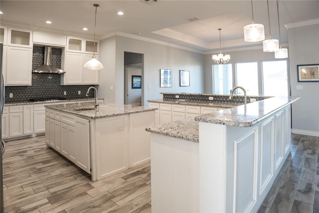 kitchen featuring wall chimney range hood, white cabinets, sink, and an island with sink