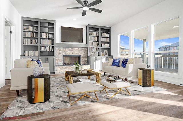 living area with wood finished floors, a ceiling fan, built in shelves, and a stone fireplace