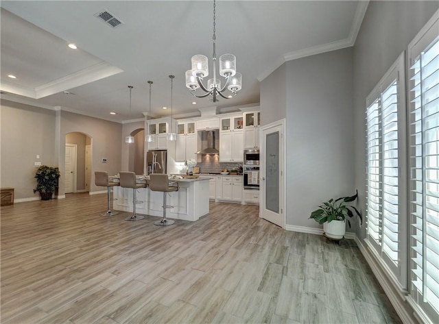 kitchen with visible vents, a kitchen island with sink, arched walkways, a breakfast bar area, and wall chimney range hood