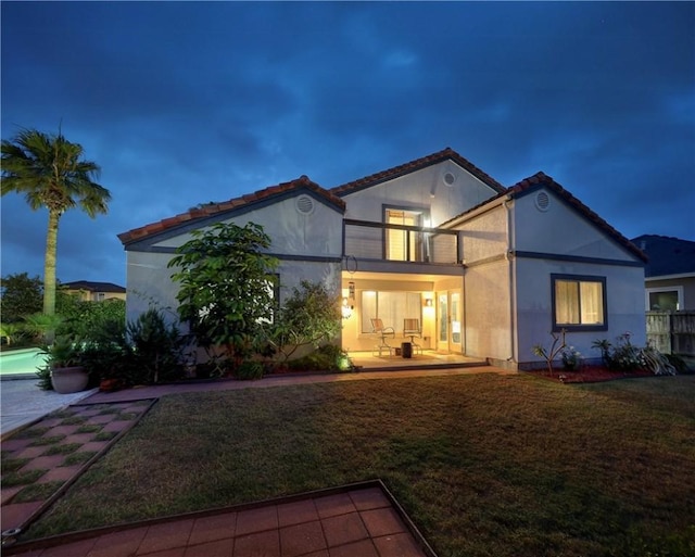 back of house at night with a lawn, a patio area, a tile roof, and a balcony