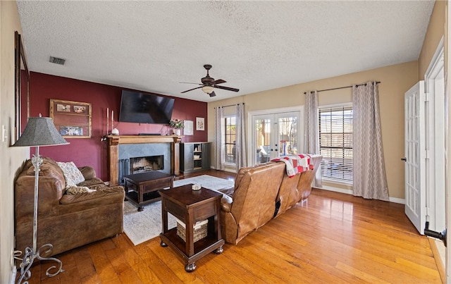 living room with a fireplace, light hardwood / wood-style flooring, ceiling fan, a textured ceiling, and french doors