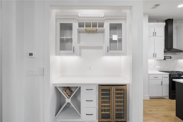 bar featuring black gas range oven, light wood-type flooring, wine cooler, wall chimney exhaust hood, and white cabinets