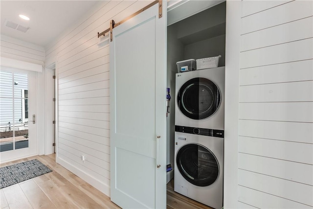 laundry area with wooden walls, stacked washer / dryer, a barn door, and light hardwood / wood-style floors