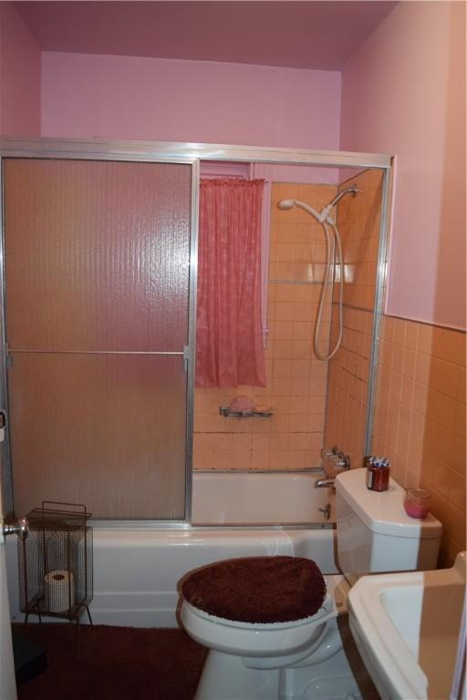 full bathroom with tile walls, toilet, a wainscoted wall, and bath / shower combo with glass door