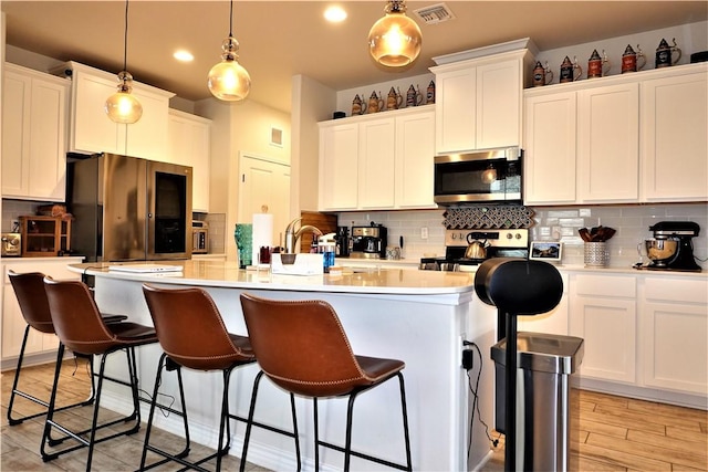 kitchen with a kitchen island with sink, hanging light fixtures, stainless steel appliances, white cabinets, and decorative backsplash