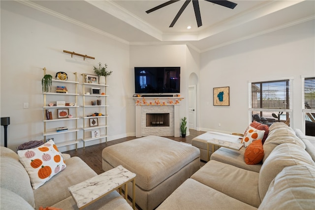 living room featuring arched walkways, a fireplace, wood finished floors, a tray ceiling, and crown molding