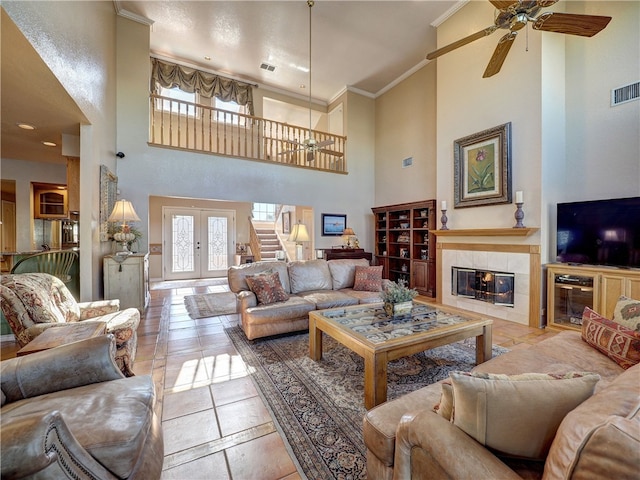 living room with crown molding, ceiling fan, a towering ceiling, a tiled fireplace, and french doors