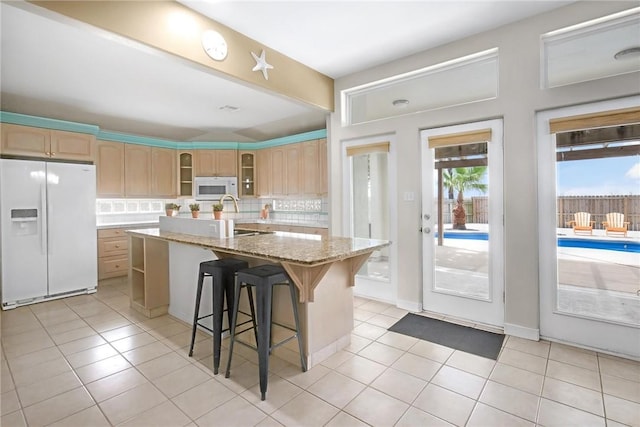 kitchen featuring white appliances, an island with sink, glass insert cabinets, backsplash, and light brown cabinetry