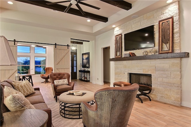 living room featuring light hardwood / wood-style floors, beamed ceiling, ceiling fan, a stone fireplace, and a barn door