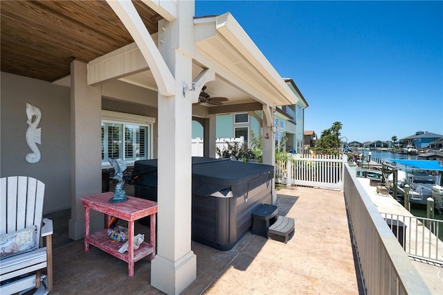 view of patio with a hot tub, a water view, a ceiling fan, and fence