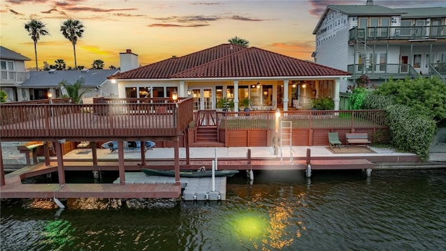 back of property at dusk with a tiled roof, a chimney, and a deck with water view