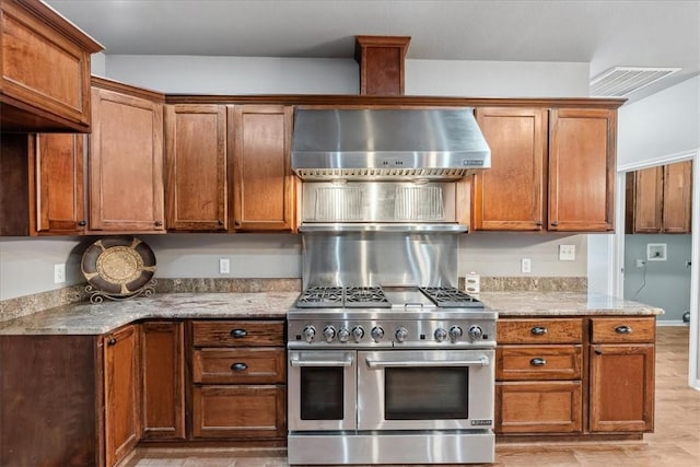 kitchen featuring visible vents, range with two ovens, wall chimney exhaust hood, light stone counters, and brown cabinets