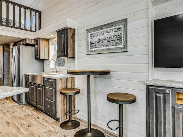 kitchen with light wood-style floors, freestanding refrigerator, a sink, wooden walls, and light stone countertops