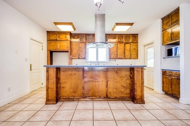 kitchen featuring baseboards, light tile patterned floors, brown cabinetry, island range hood, and a sink