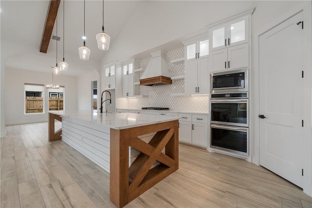 kitchen with custom exhaust hood, decorative light fixtures, built in microwave, a kitchen island with sink, and white cabinets