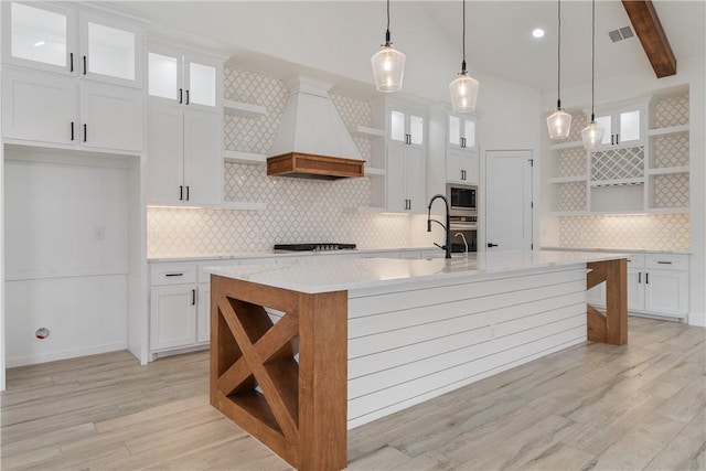 kitchen featuring a kitchen island with sink, appliances with stainless steel finishes, hanging light fixtures, custom range hood, and white cabinetry