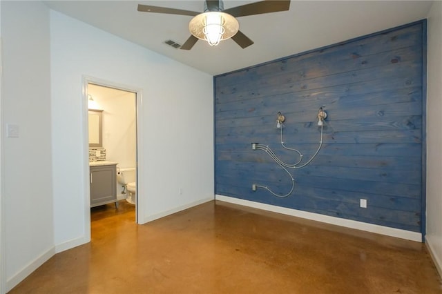 unfurnished bedroom with ceiling fan, ensuite bathroom, wooden walls, and concrete floors